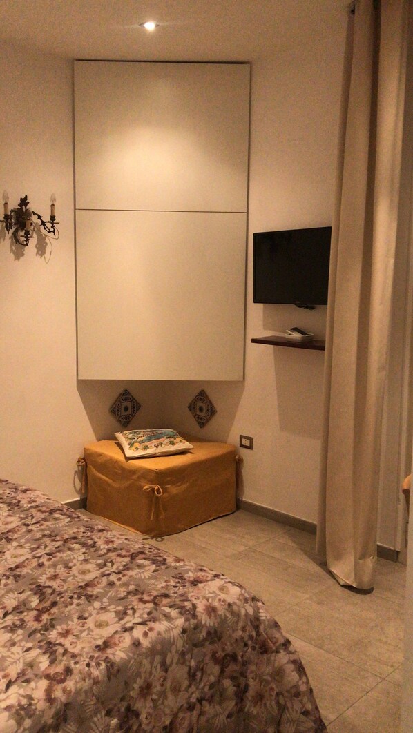 A bedroom with a bed, a television mounted on the wall, a small table with a cushion, and a curtain. - International Naples (Naples)