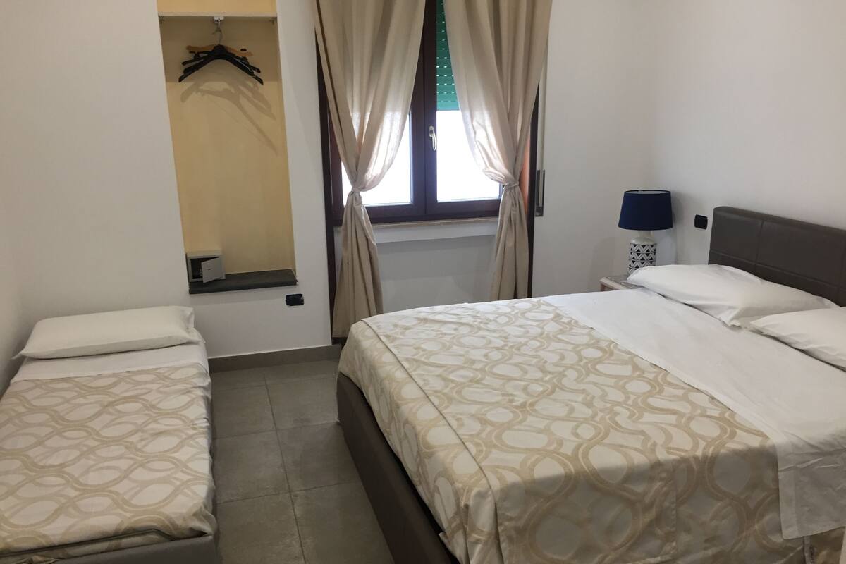 A bedroom with a bed, a smaller bed, a nightstand with a lamp, a window with curtains, and a wardrobe with hanging clothes.