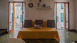 A room with a yellow bed, two doors leading to a balcony, a wall clock, and a shelf with small items. - El Nin Apartment with Terrace & AC (Vernazza)