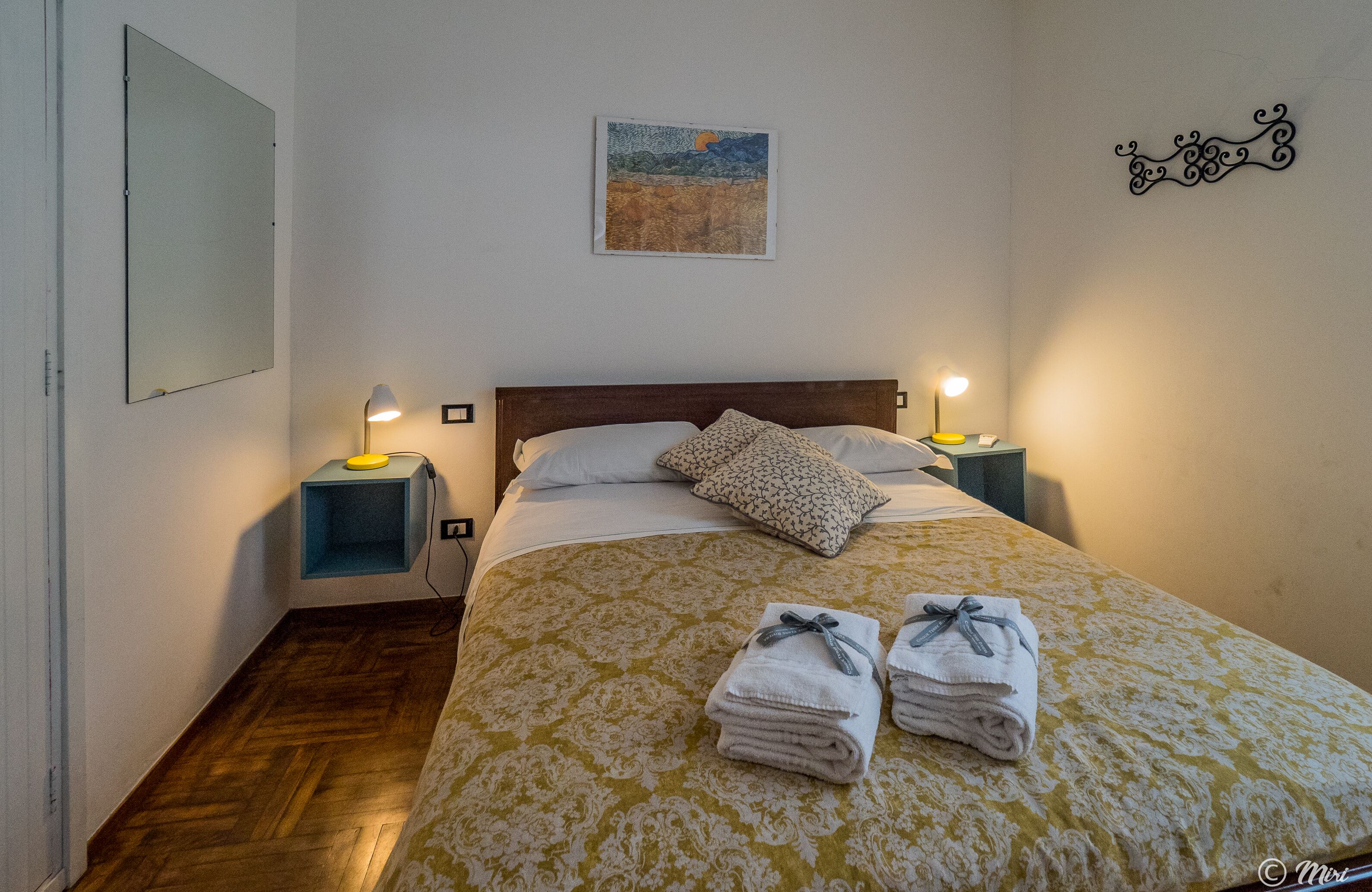 A neatly made bed with a patterned bedspread, two bedside lamps, a wall-mounted artwork, and a decorative wall piece.