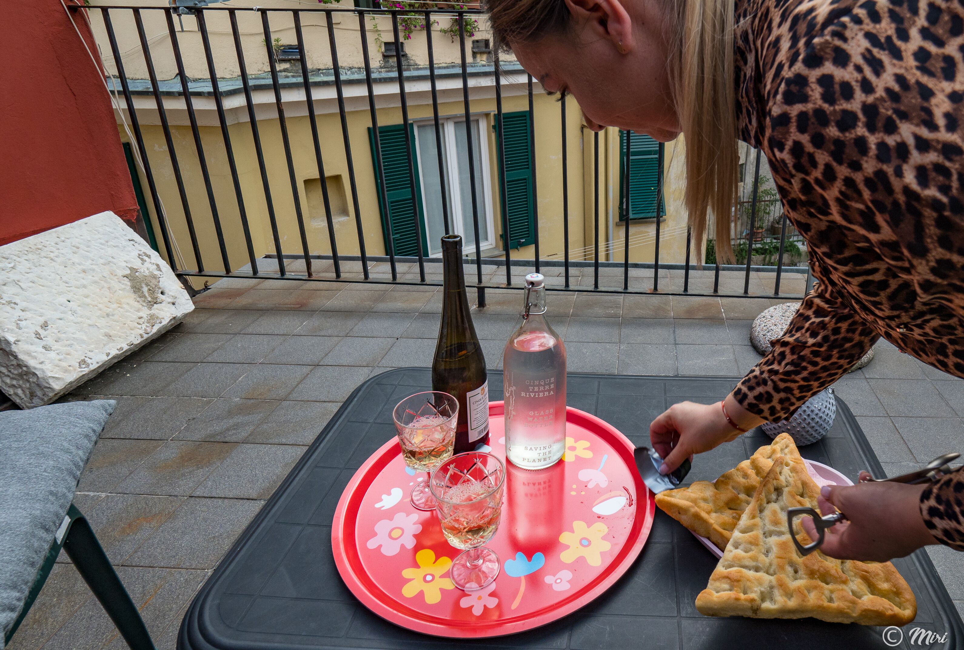 Een tafel met een dienblad, een fles, glaasjes en een driehoekig gebakje op een balkon.