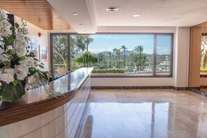 Zona de recepción de un hotel con suelo de mármol, mostrador de recepción y un amplio ventanal que ofrece vistas a palmeras y montañas.