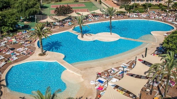 A large, circular swimming pool with multiple sunbathing areas and palm trees. - Aparthotel Club Cala Romaní (Cales de Mallorca)