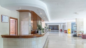 A modern hotel lobby with a curved reception desk, a large plant, and a glass door leading to the entrance. - Aparthotel Club Cala Romaní (Cales de Mallorca)