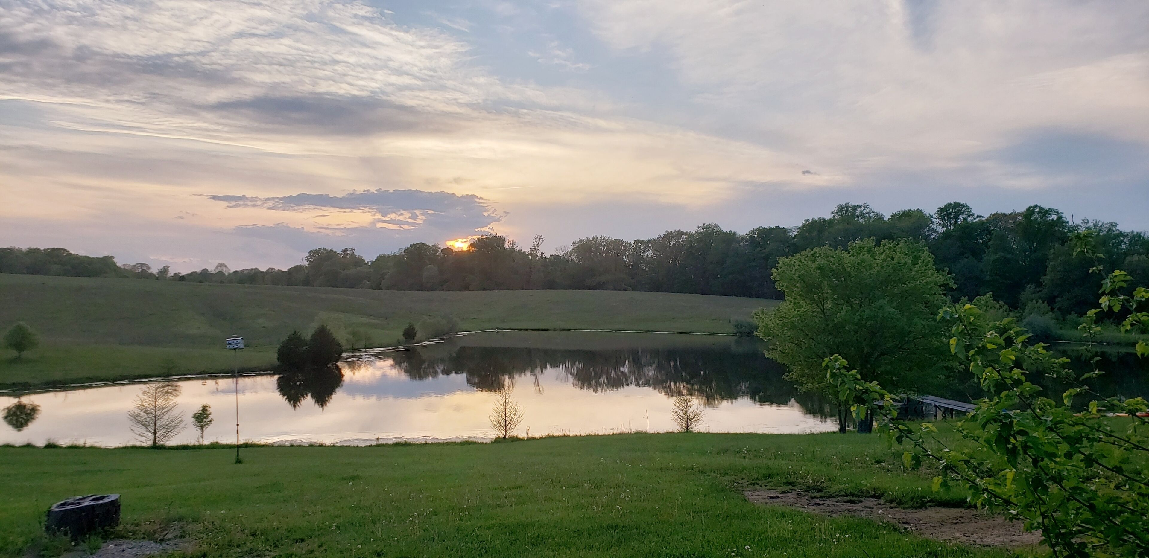 A serene landscape with a pond reflecting the sky, surrounded by green fields and trees.