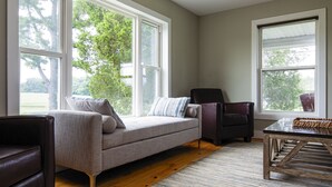 A spacious living room with a grey sofa, a dark armchair, and a wooden coffee table. Large windows provide natural light and a view of trees. - Wine Country Farmhouse Private Lake! kayaks Fire Pit! On The Wine Trail! (Cobden)