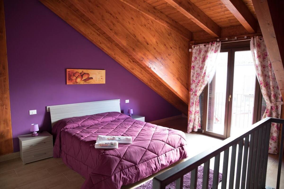 Un dormitorio con pared morada, techo de madera, cama con cubrecama morado, mesita de noche con lámpara y ventana con cortinas florales.