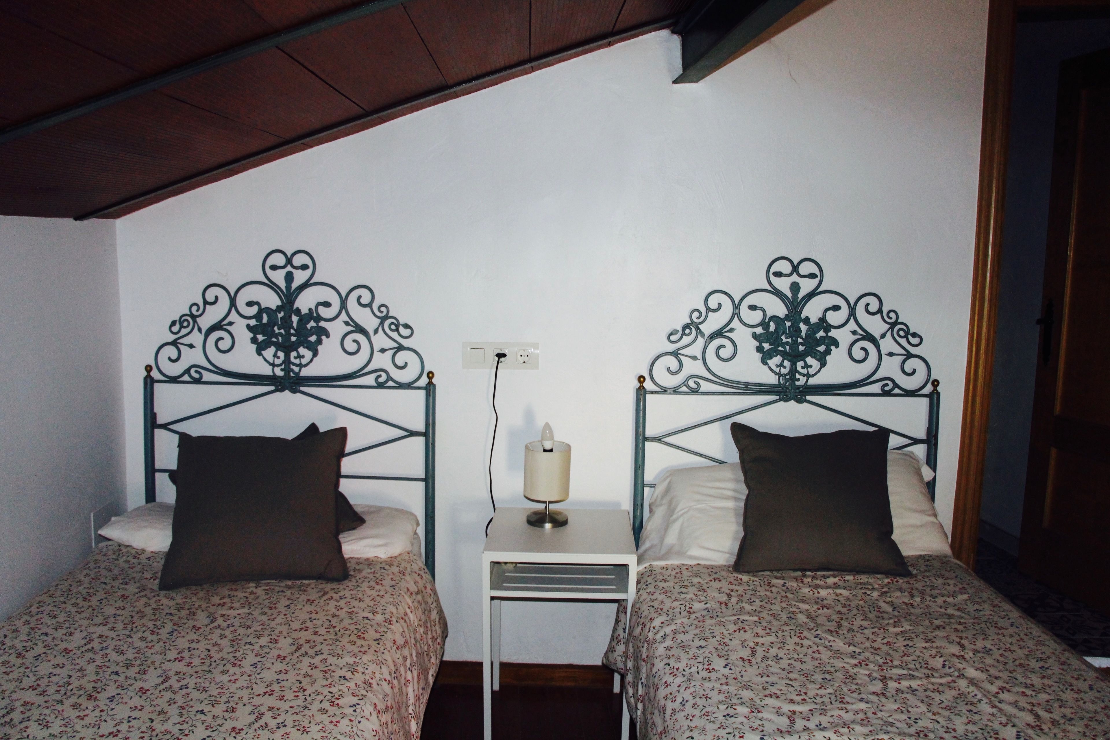 Una habitación con dos camas, cada una con una colcha estampada y almohadas oscuras. Hay una mesita de noche blanca pequeña entre las camas con una lámpara encima.