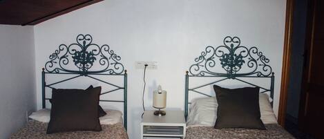 Una habitación con dos camas, cada una con una colcha estampada y almohadas oscuras. Hay una mesita de noche blanca pequeña entre las camas con una lámpara encima.