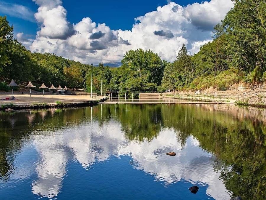 Ein ruhiger Poolbereich im Freien mit blauem Himmel, umgeben von Bäumen und einer Brücke.