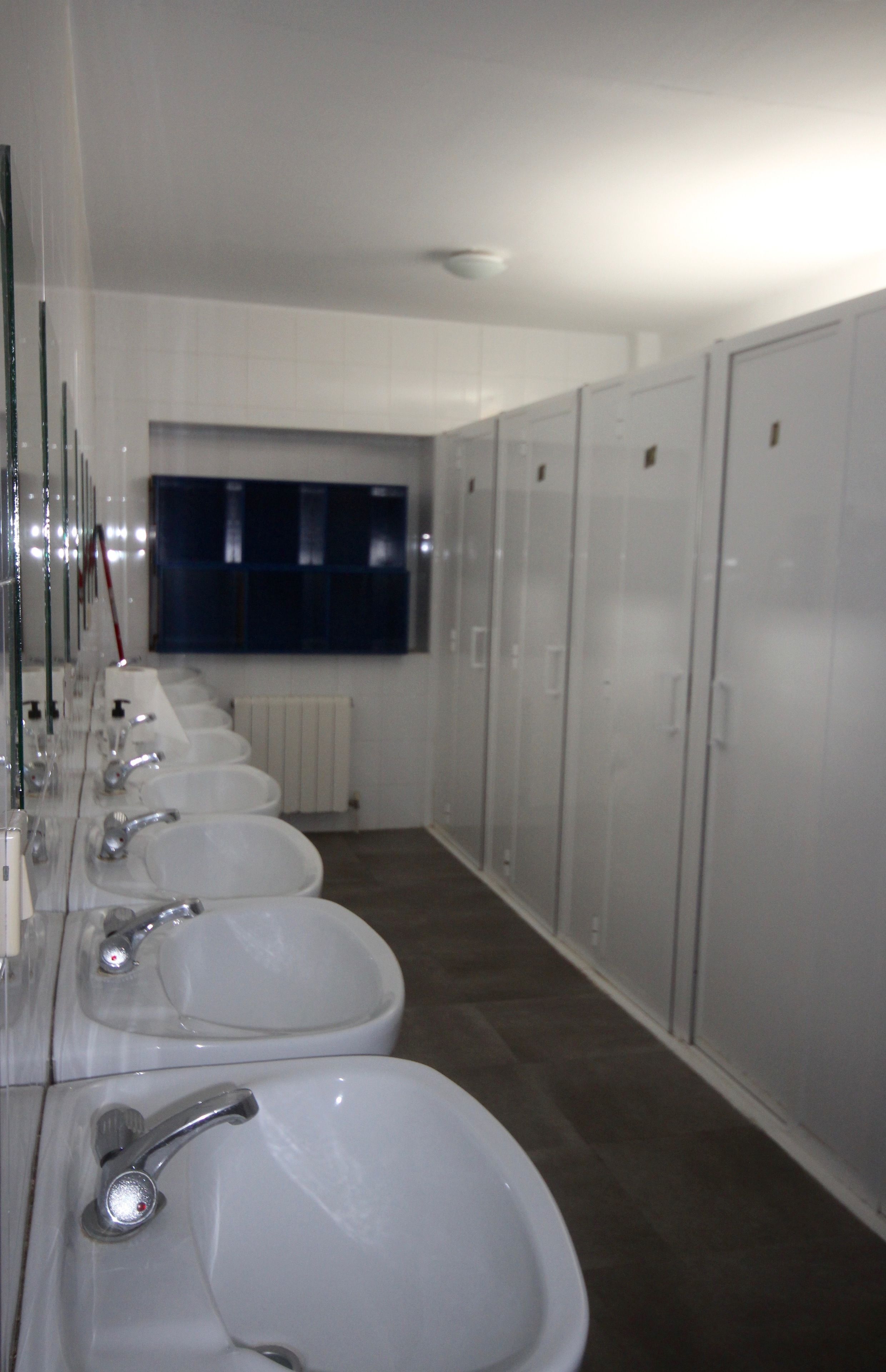 A row of white sinks with mirrors, a window, and lockers along the wall.