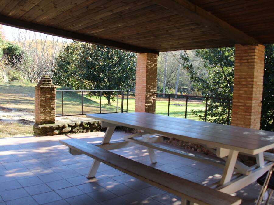 Un patio al aire libre con una mesa de picnic y bancos, rodeado por un muro de ladrillo y una barandilla de metal que da a un área verde con árboles.