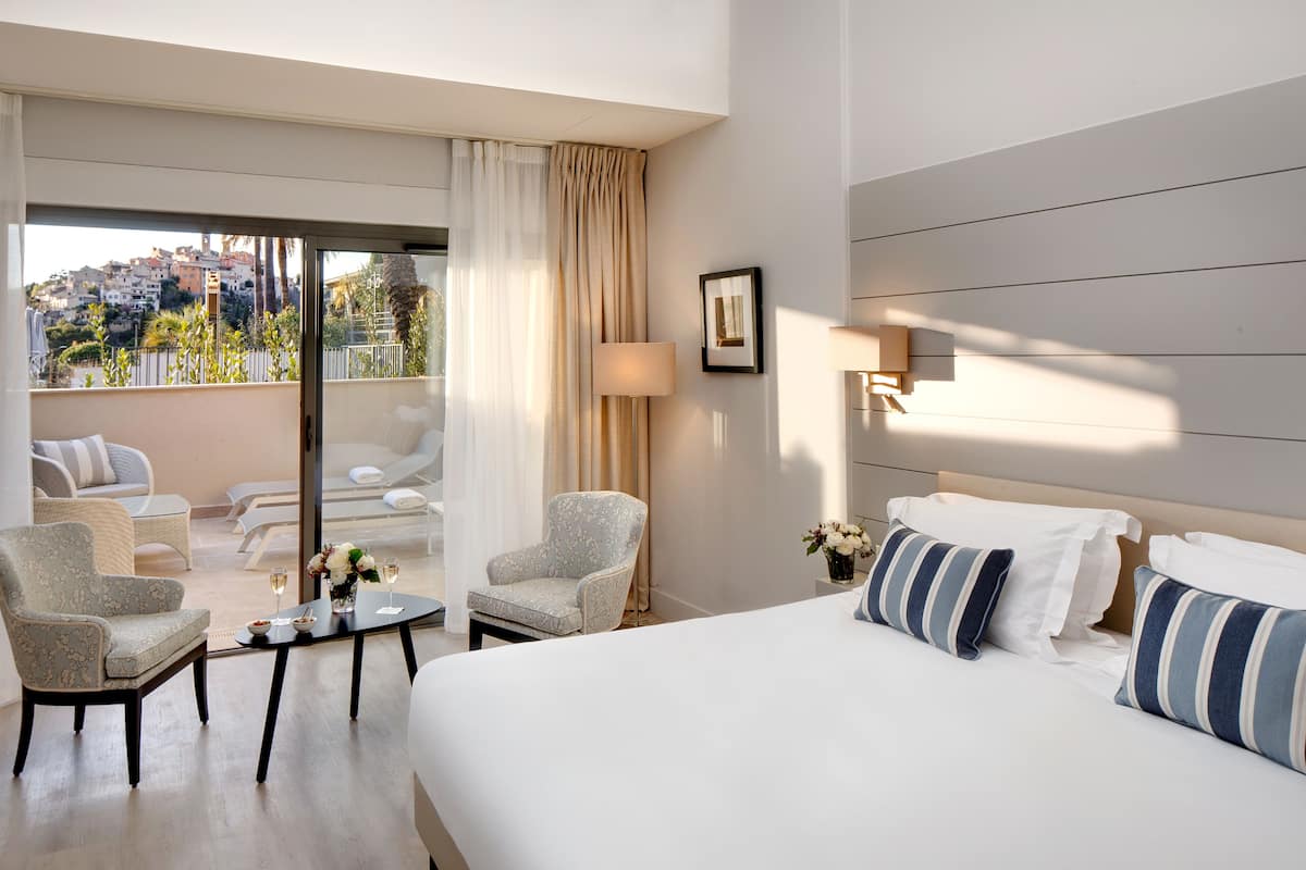 A hotel room with a large bed, two armchairs, a small table with a vase of flowers, and a view of the city through large windows.