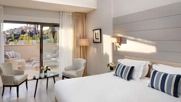 Quarto de hotel com uma cama grande, duas poltronas, uma mesinha de centro com um vaso de flores e vista para a cidade através de amplas janelas.