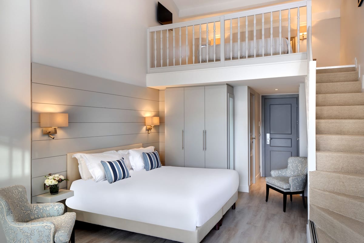 A bedroom with a large bed, a nightstand, a chair, a wardrobe, and a staircase leading to an upper level.