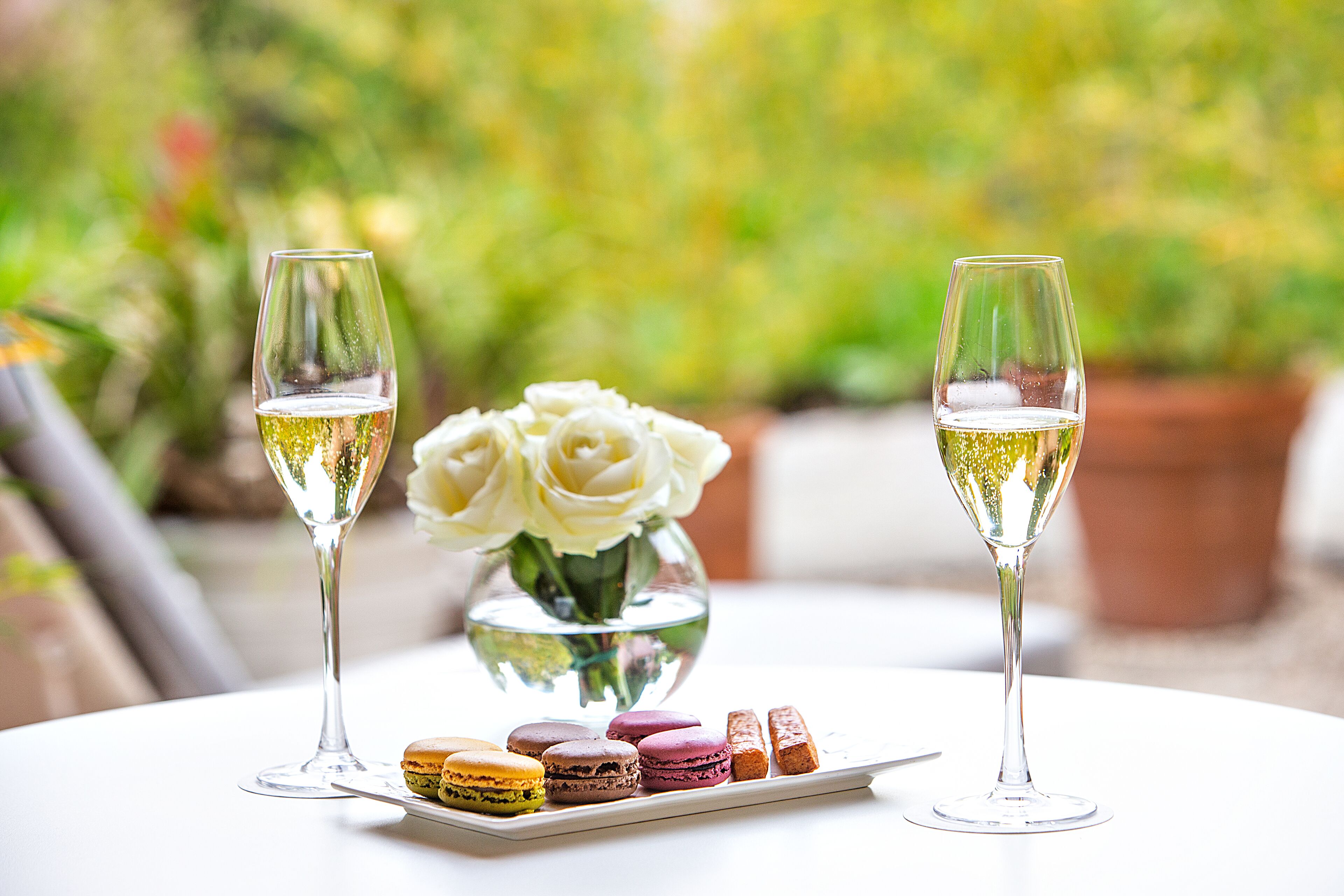 Two glasses of white wine on a table with macarons and a vase of white roses.