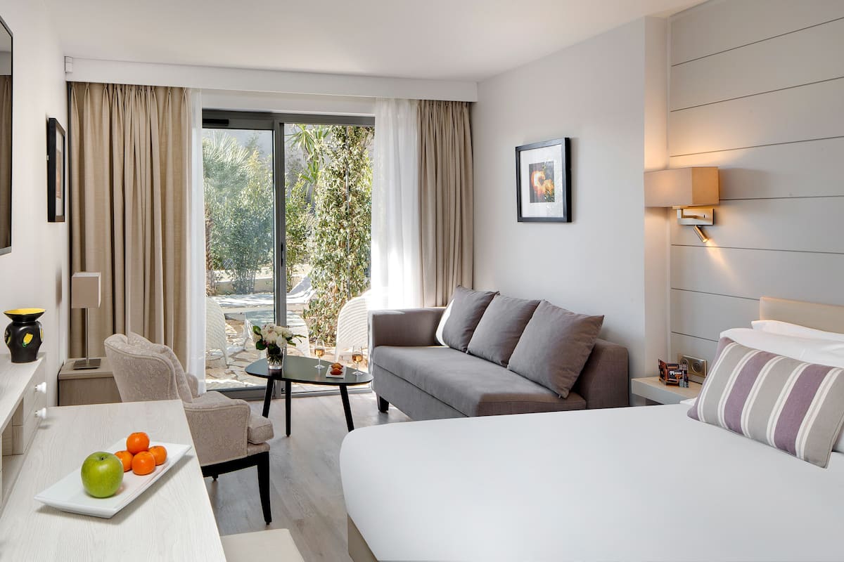 A modern hotel room with a sofa, a bed, a dining table with fruit, and a view of greenery outside.