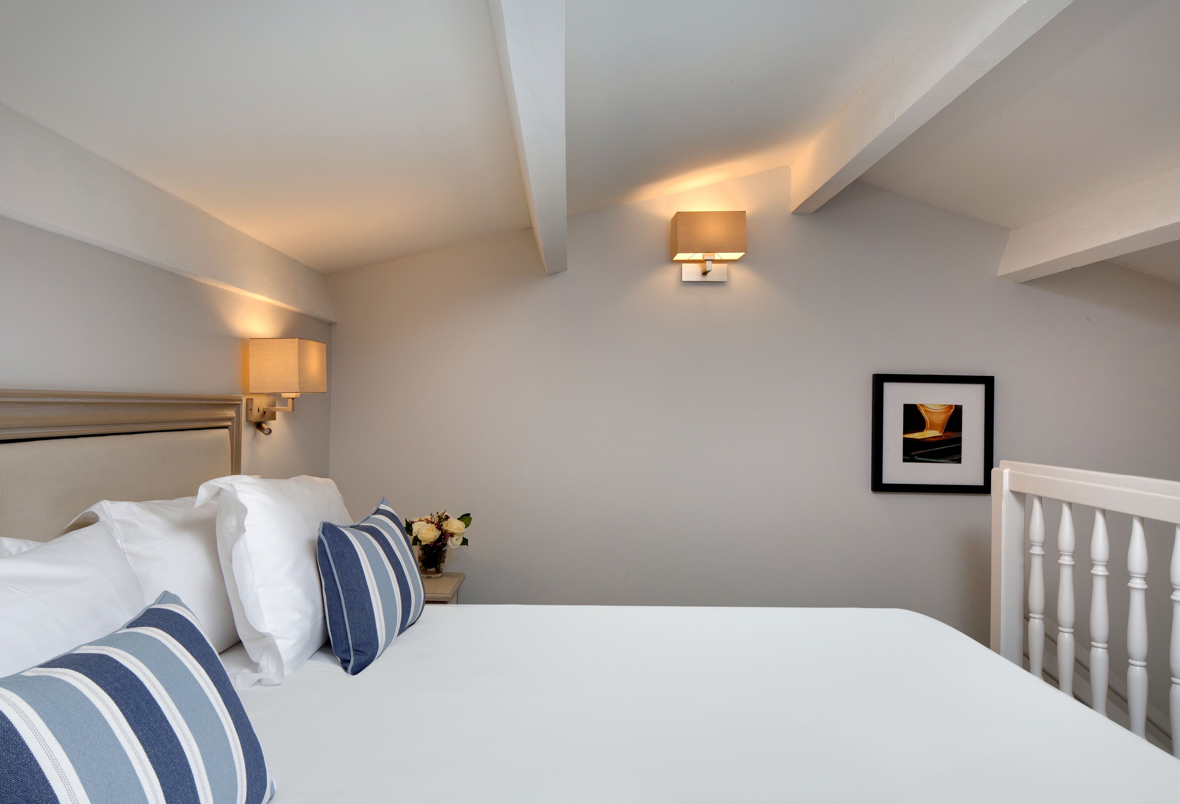 Un dormitorio ordenado con cama, almohadas, una mesita de noche con un florero y un cuadro en la pared.