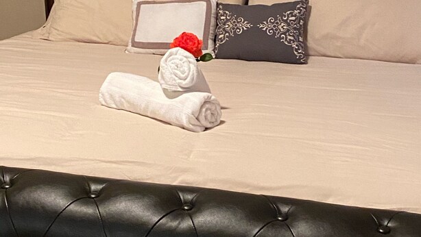 Ein Bett mit einem geblatteten Kopfteil, zwei Kissen, einem zusammengerollten Handtuch und einer roten Rose.
