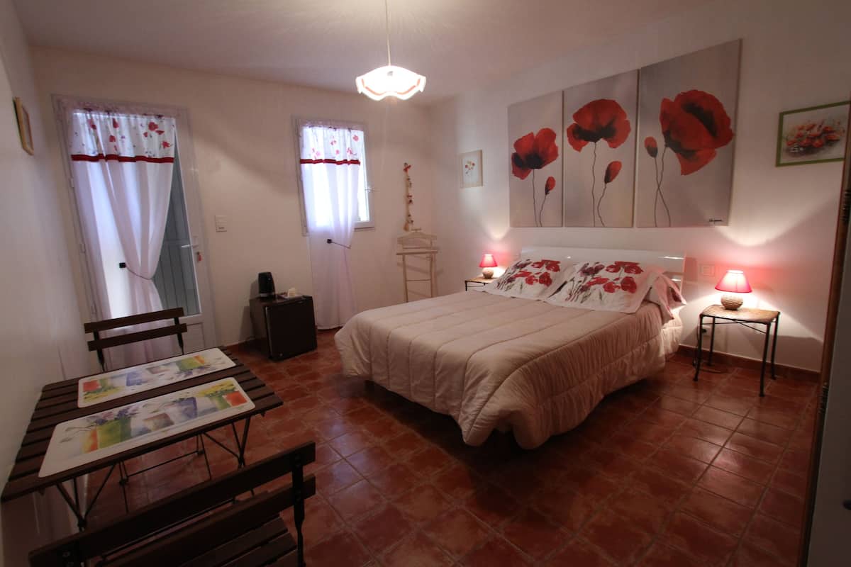 A bedroom with a bed, bedside tables, a desk with a chair, a window with curtains, and a wall with floral artwork.