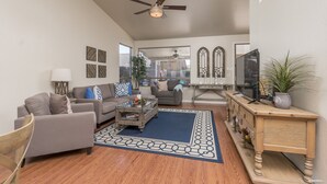 A spacious living room with a ceiling fan, a large window, a wooden coffee table, and a blue patterned rug.