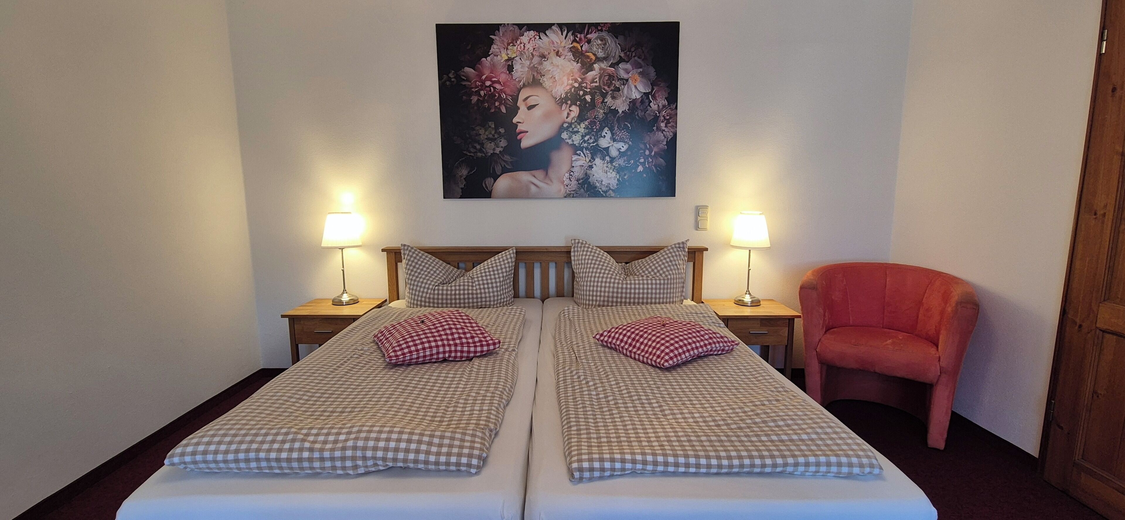Ein Doppelbett mit karierter Bettwäsche, zwei Nachttischlampen, ein roter Sessel und ein Blumenbild an der Wand.