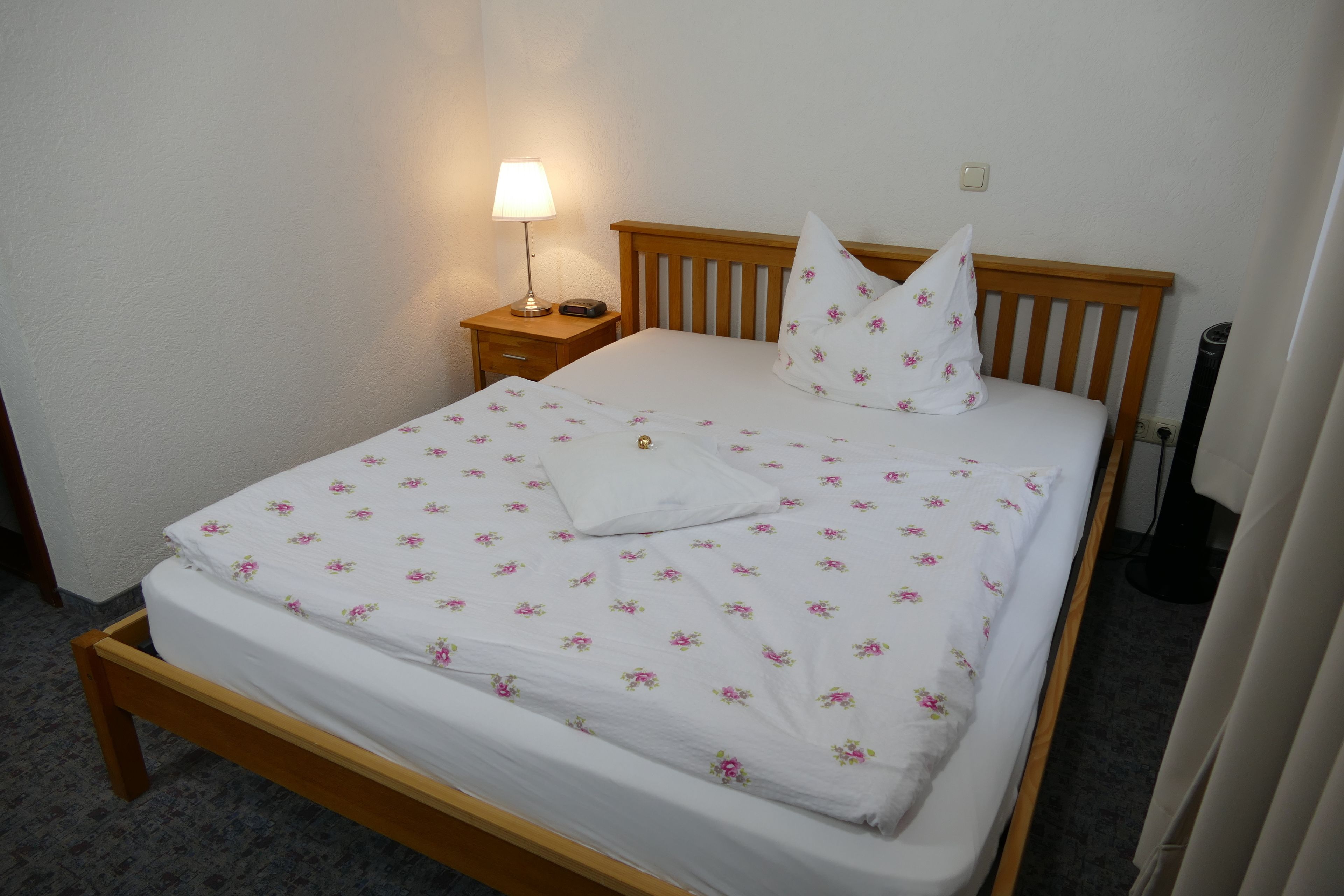 A neatly made bed with a floral bedspread, a wooden headboard, a bedside lamp, and a small bedside table.