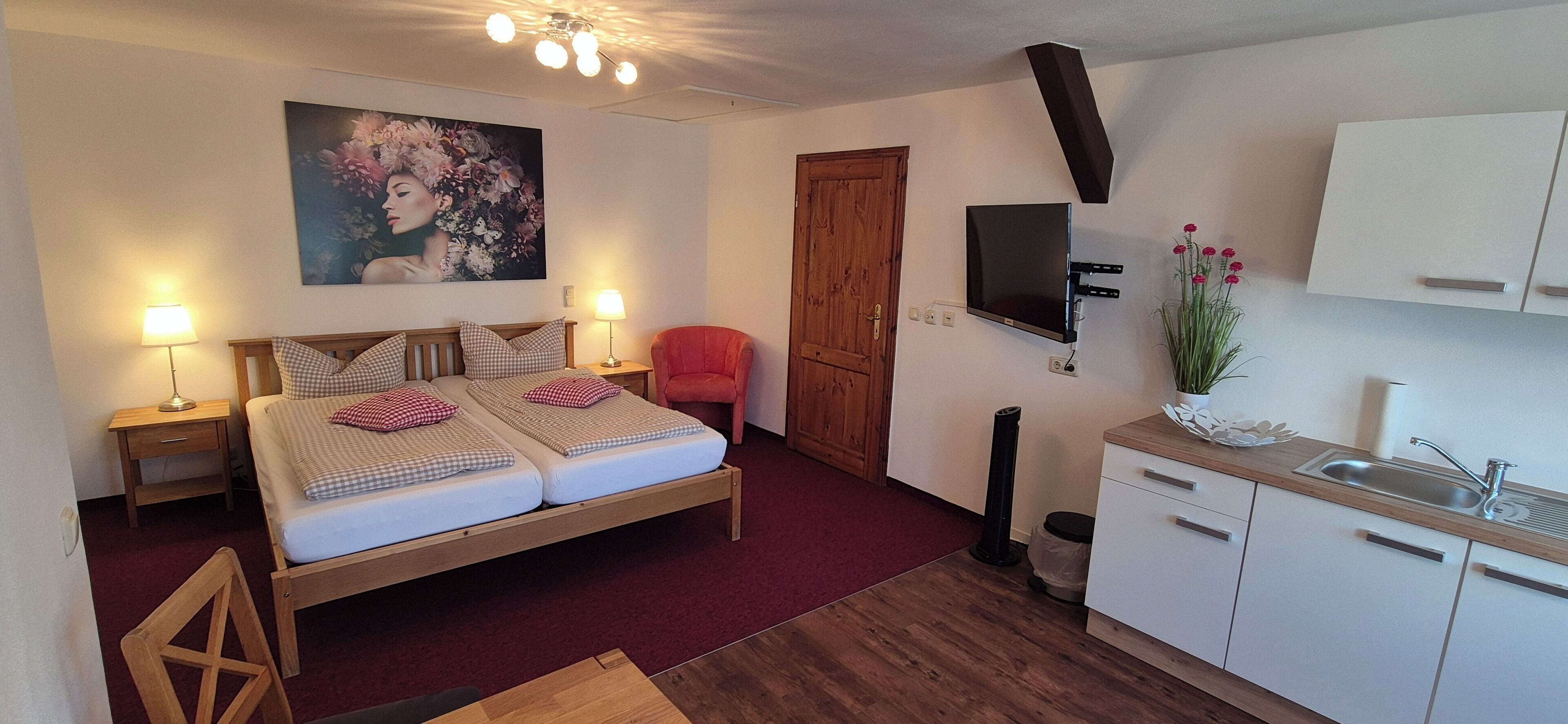 Ein Schlafzimmer mit einem Doppelbett, Nachttischlampen, einem Holzstuhl, einem roten Sessel, einem Holzschrank, einem an der Wand befestigten Fernseher und einer kleinen Küchenzeile mit Spüle und Schränken.
