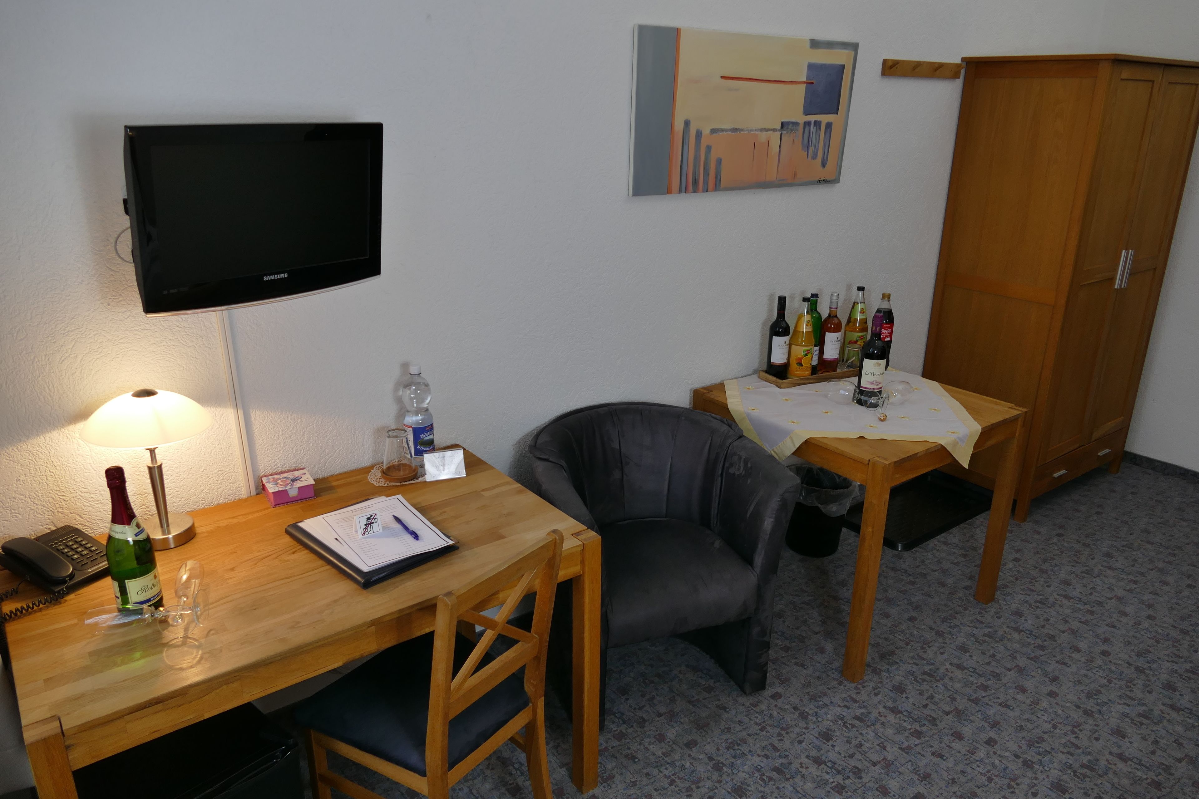 A hotel room with a wooden desk, a chair, a television mounted on the wall, a cabinet, and a table with bottles.
