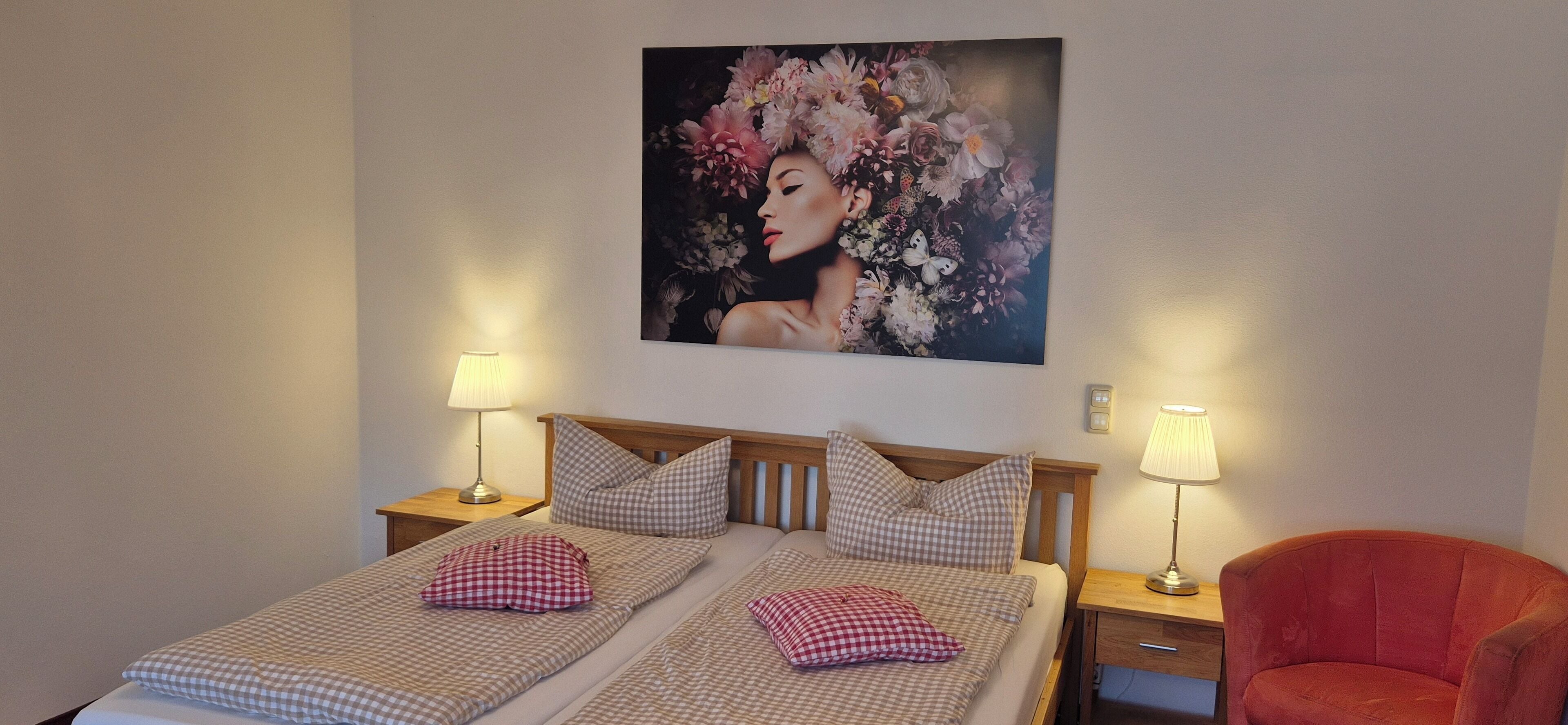 A double bed with checkered bedding, two bedside tables with lamps, a red armchair, and a floral artwork on the wall.