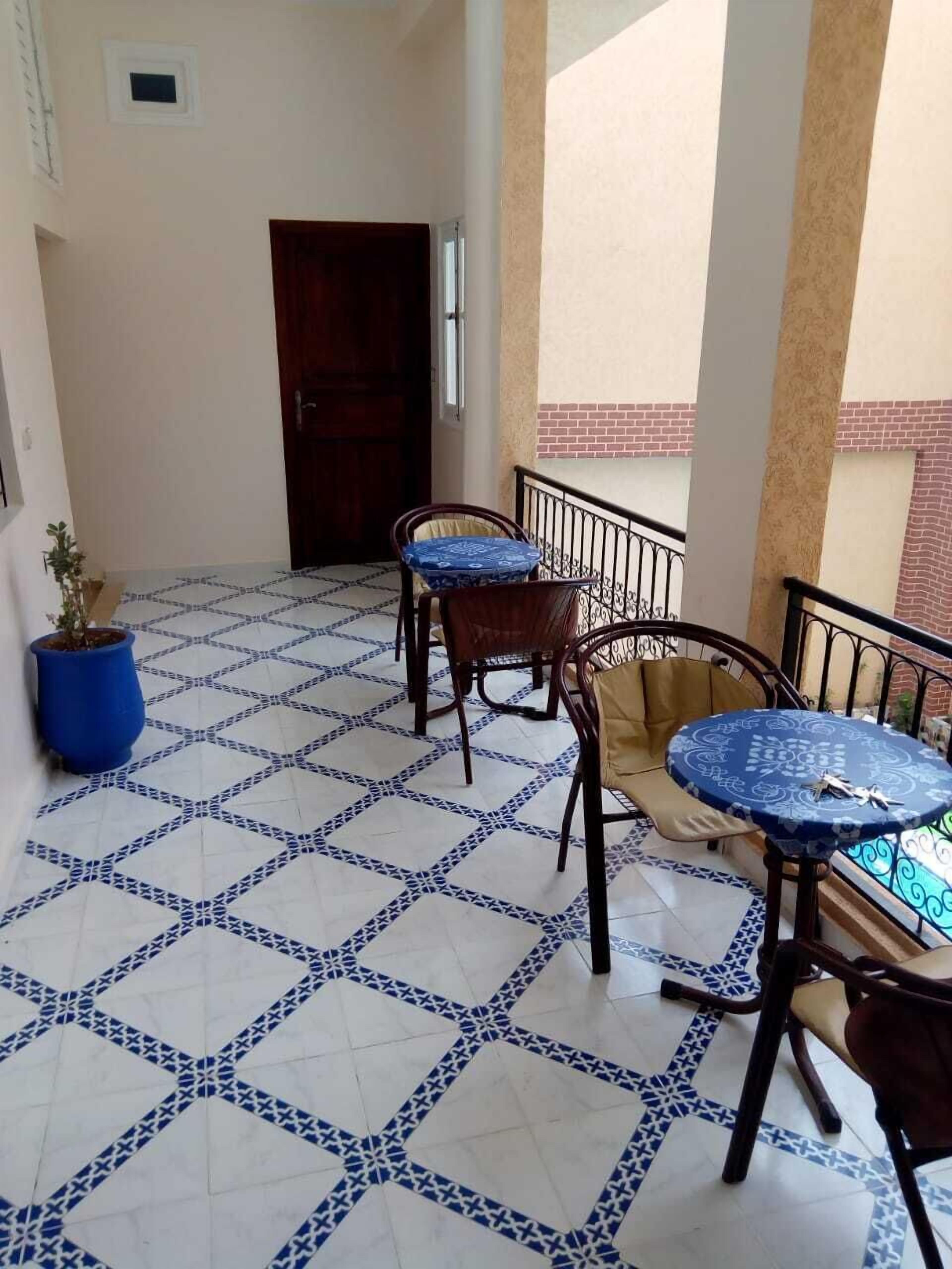 A balcony with patterned floor tiles, two wooden chairs, a small round table, and a potted plant.