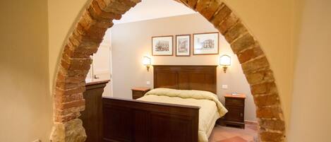 A bedroom with a brick archway, a wooden bed, a nightstand, a dresser, and three framed pictures on the wall.