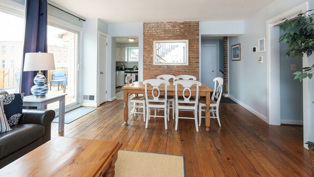 Zona pranzo con tavolo in legno e sedie bianche, parete decorata con mattoni e vista sulla cucina tramite una porta aperta.