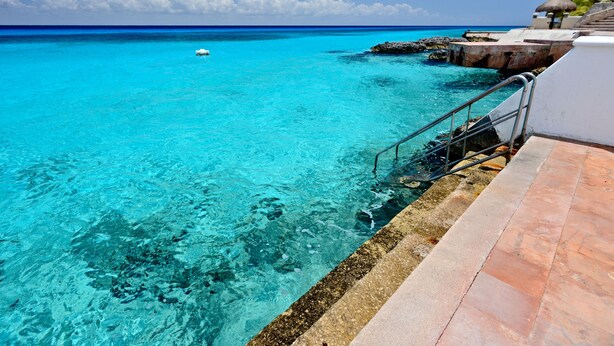 Une mer turquoise aux eaux limpides, un bateau, un escalier en pierre menant à une plage et un parasol en chaume.