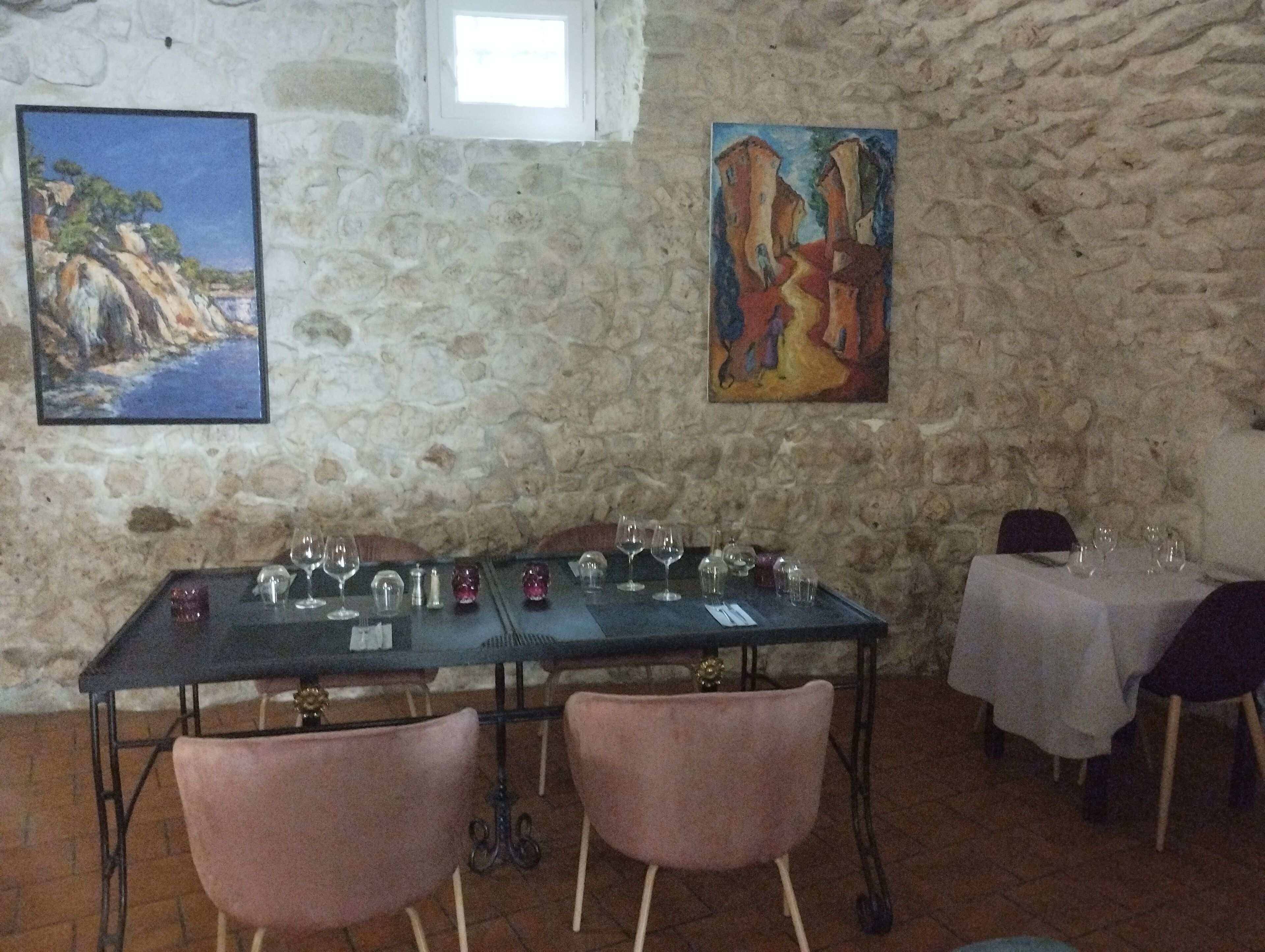 A dining area with a stone wall, two paintings, a table set with wine glasses, and pink chairs.
