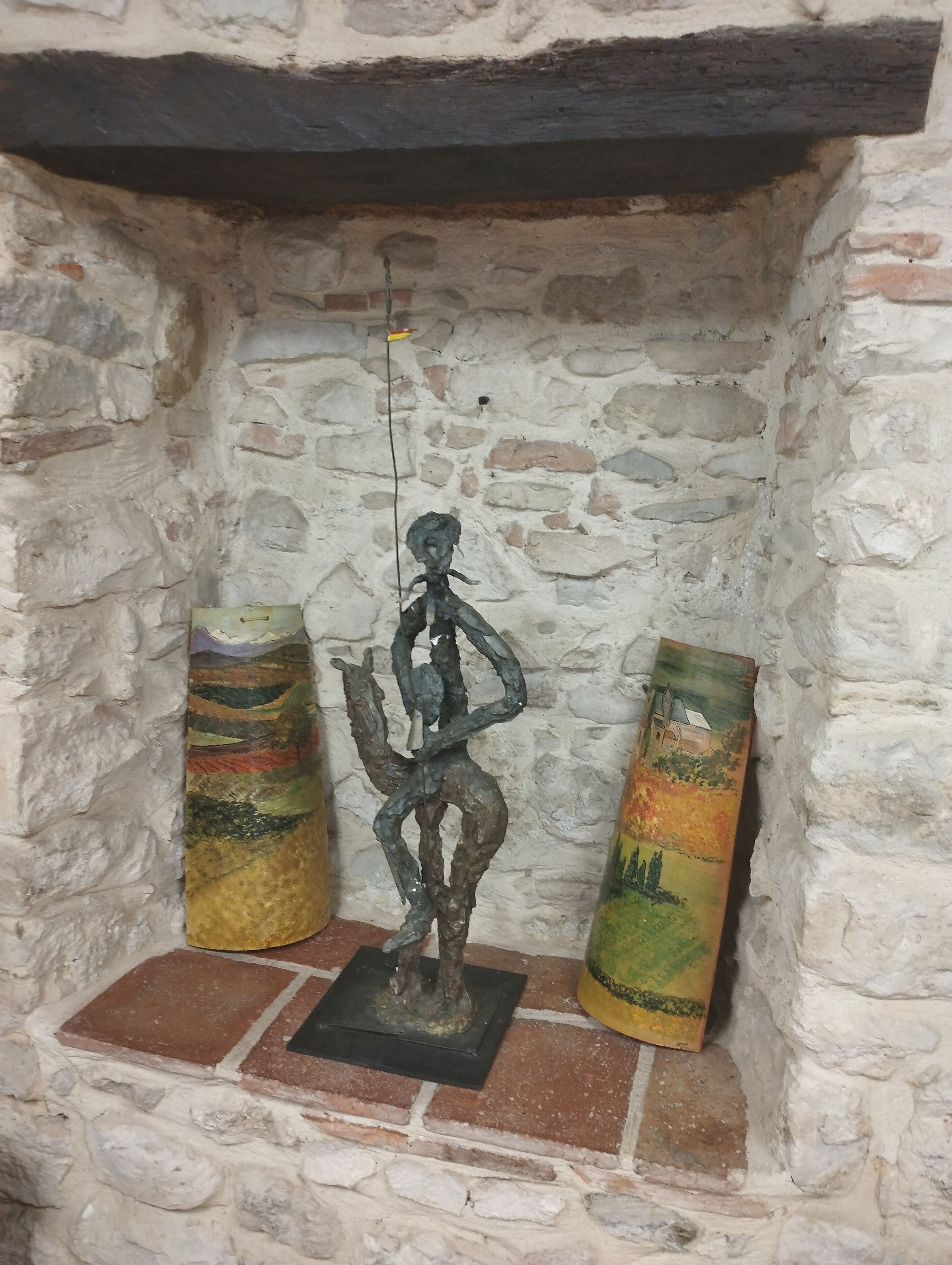 Een stenen muur met een rustieke houten plank. Op de plank staat een metalen sculptuur van een figuur die een staf vasthoudt, omringd door twee cilindervormige potten met schilderijen.