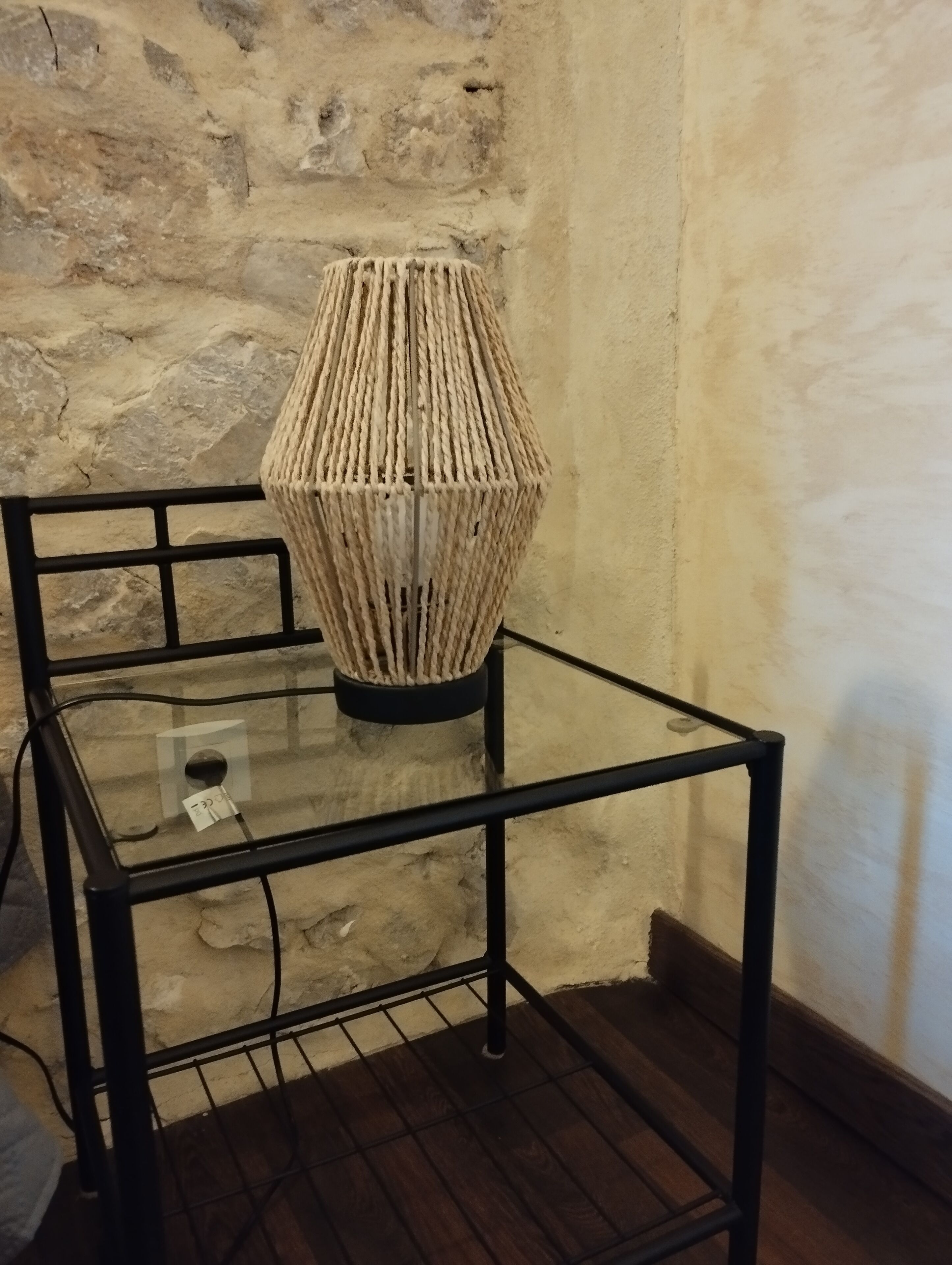 Une table à plateau en verre sur laquelle repose une lampe en osier, le tout placé devant un mur en pierre.