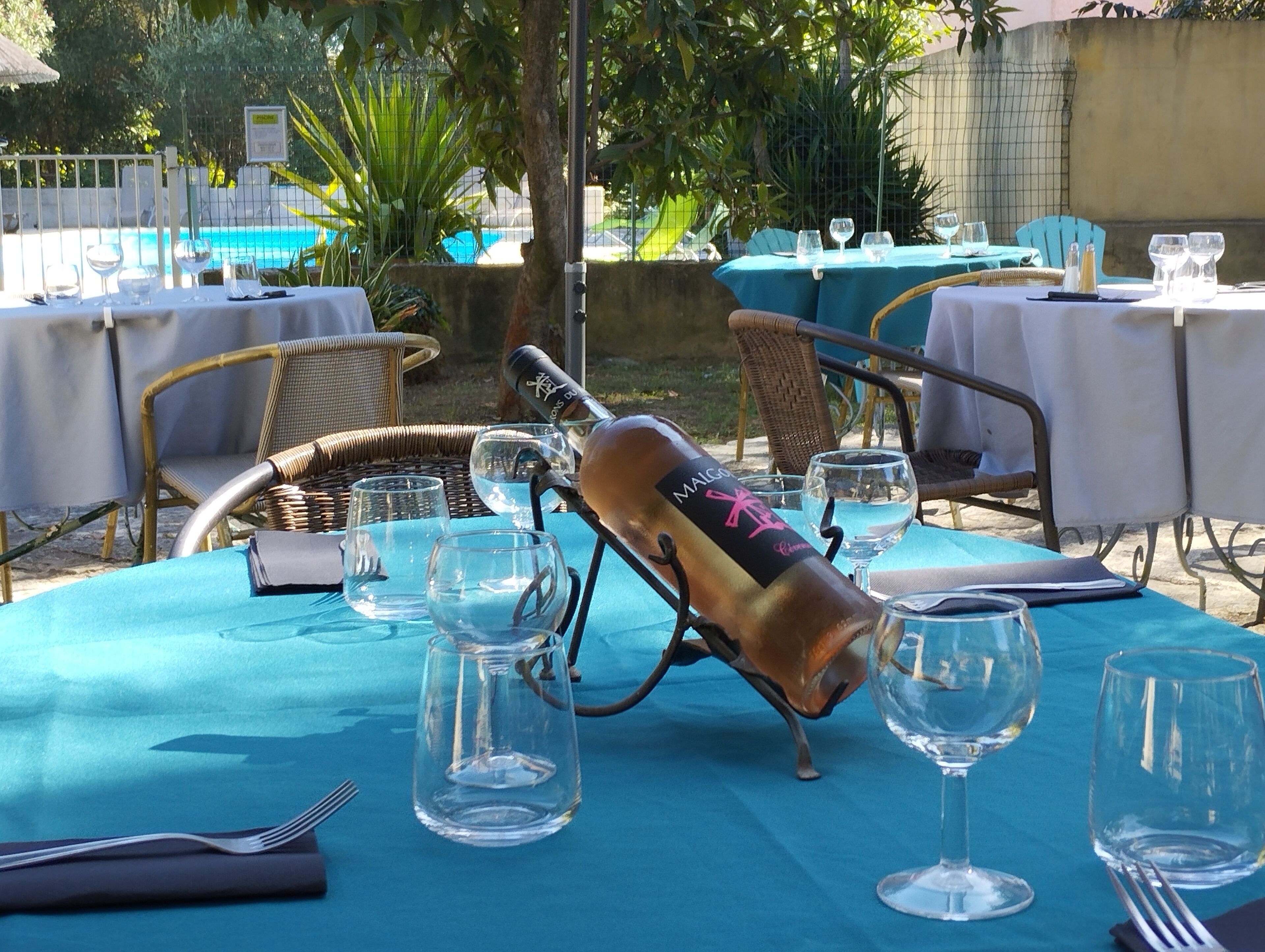 An outdoor dining area with tables set for two, featuring clear glassware and a bottle of wine.
