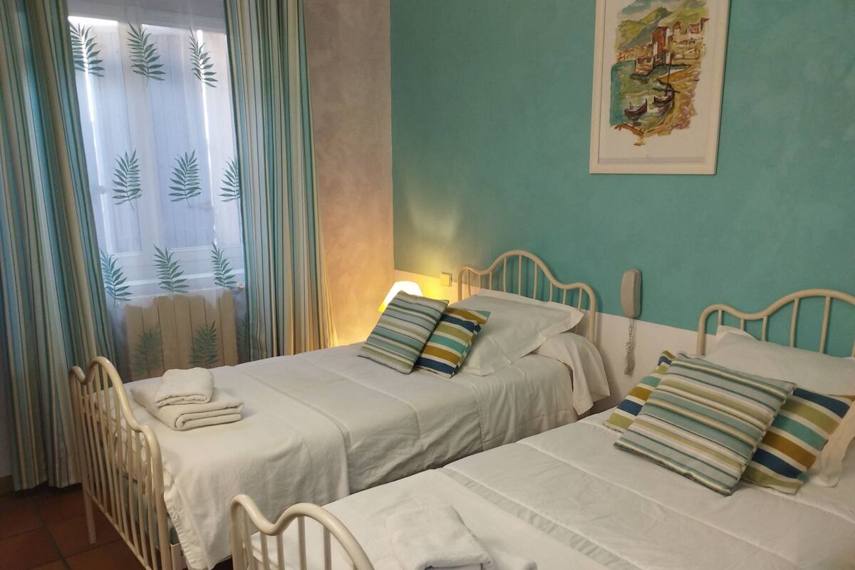 Een kamer met twee bedden, elk voorzien van witte lakens en gestreepte kussens, een nachtkastje met een lamp, en een raam met gordijnen.