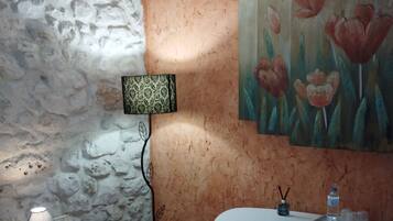 Una habitación con pared de piedra, una mesa blanca con sillas, una lámpara y un cuadro de flores.