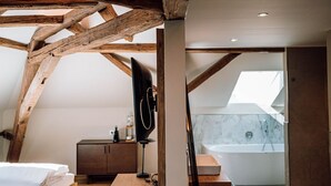 Una camera da letto con soffitto in legno, un letto, una scrivania, uno specchio e un bagno con vasca e finestra sul tetto.