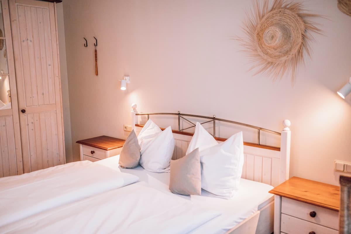 A neatly made bed with white bedding and multiple pillows, a wooden nightstand with a lamp, and a wall-mounted decorative item.