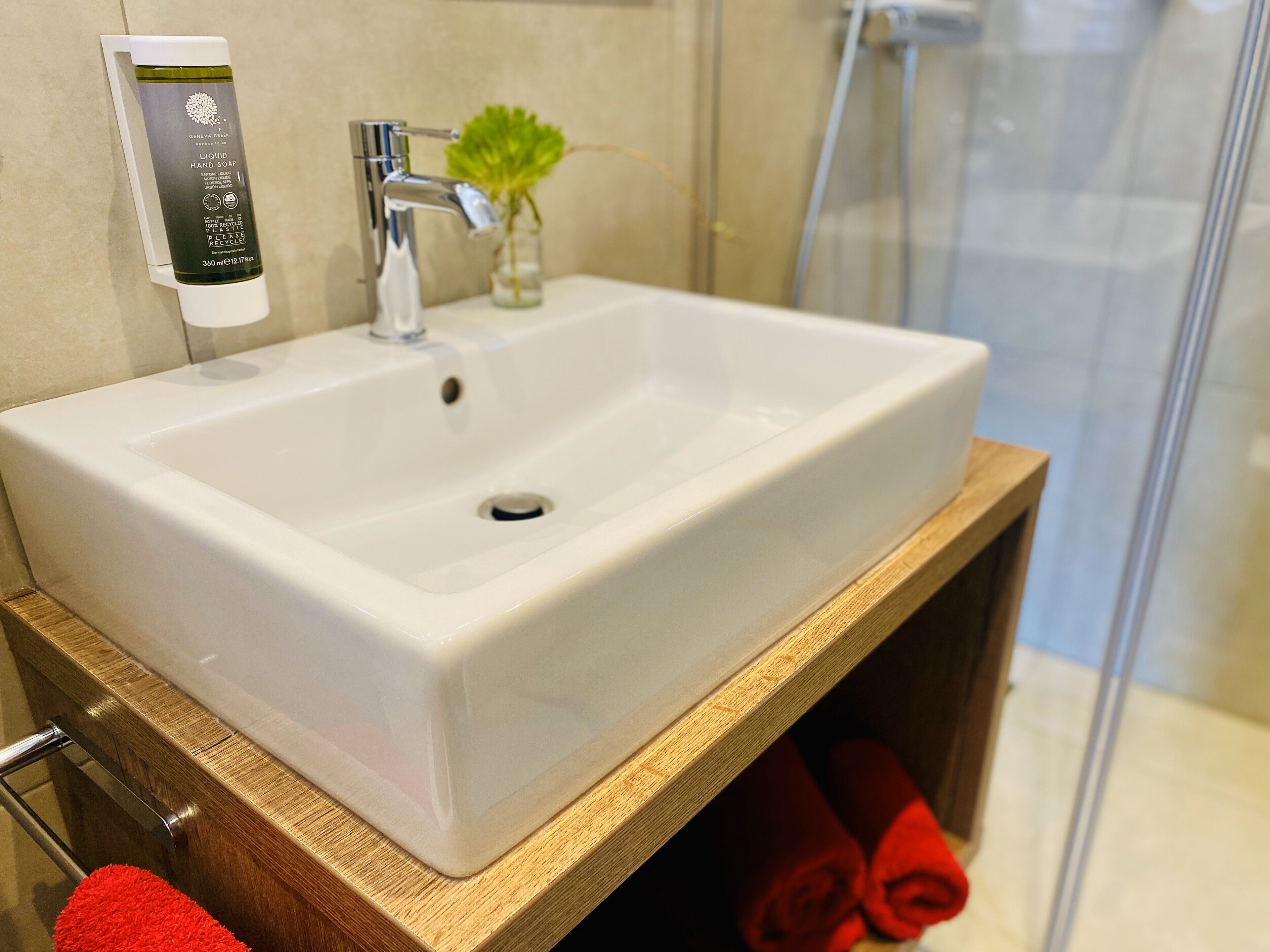 Un baño con un lavamanos blanco, un dispensador de jabón, una planta pequeña y un estante de madera con toallas.