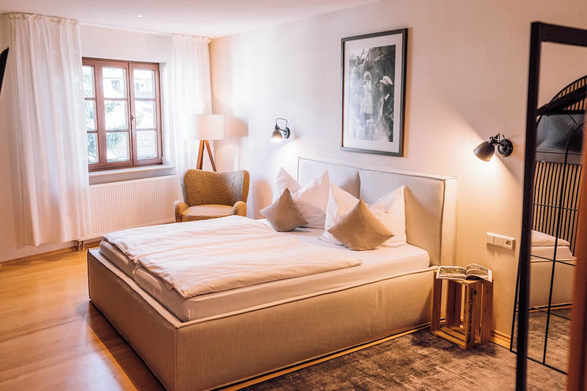 A bedroom with a large bed, a chair, a bedside table with a lamp, a framed picture on the wall, and a window with curtains.