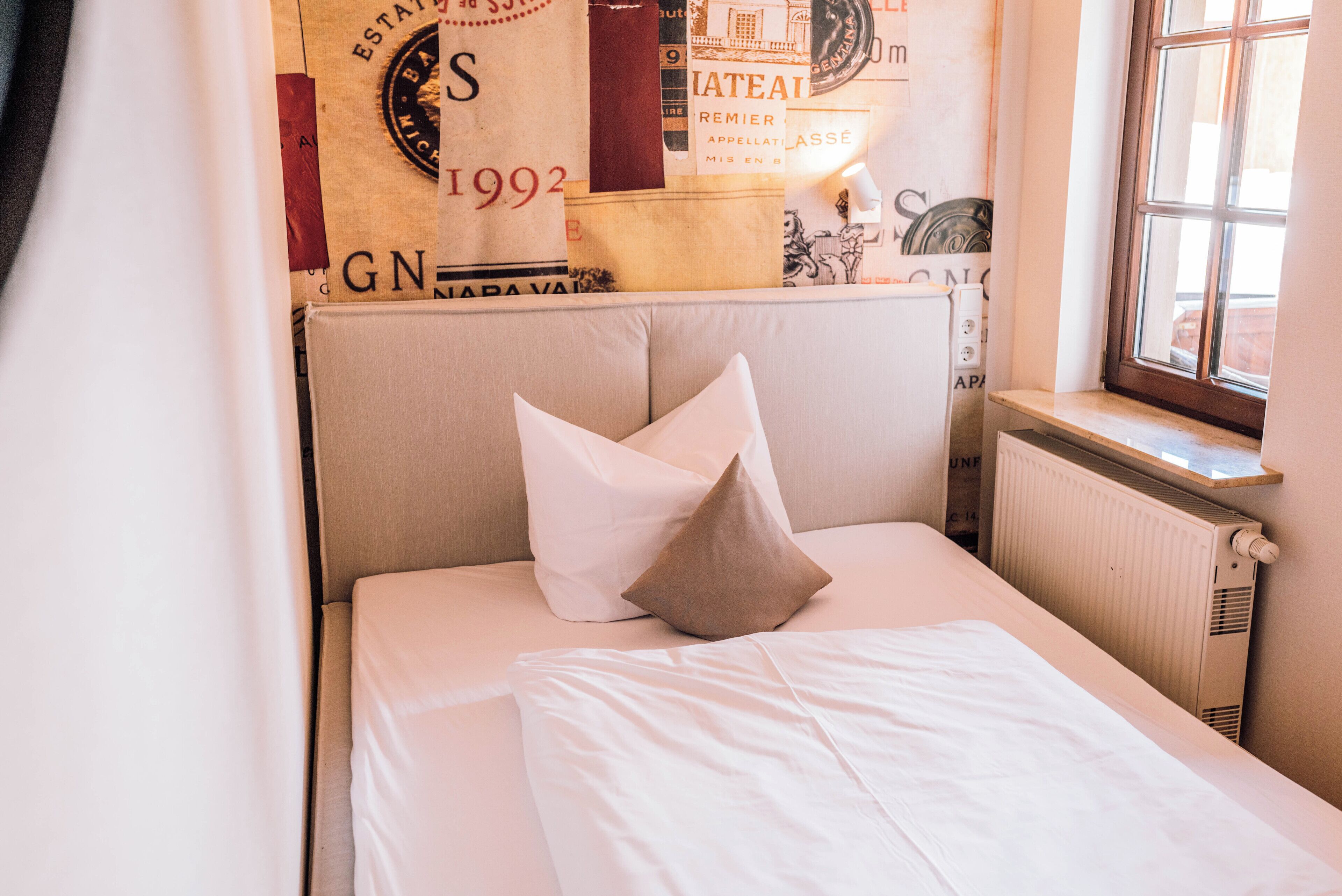 A neatly made bed with white linens and two pillows, a radiator on the wall, and a window with a view.