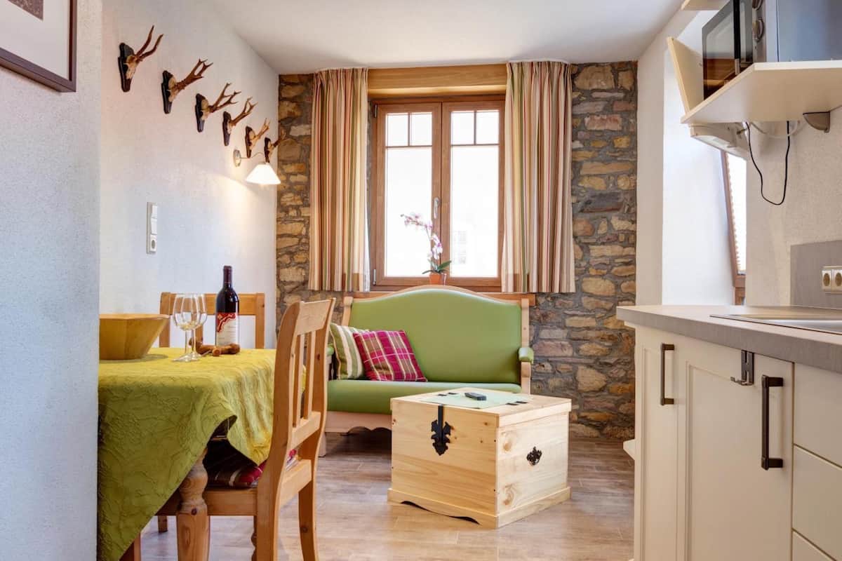 A cozy room with a wooden dining table, a green sofa, a wooden chest, and a stone wall.