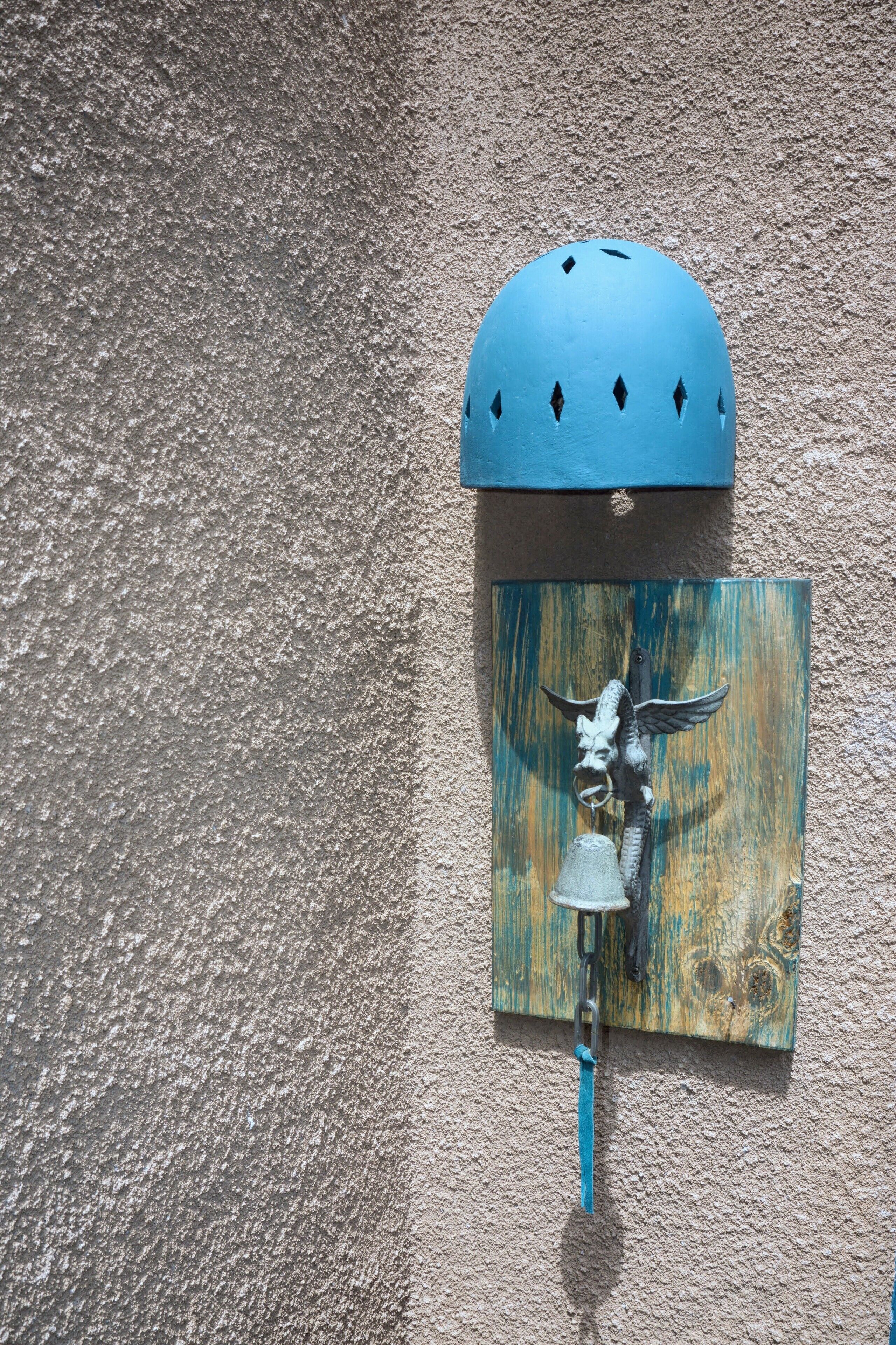 A blue bell mounted on a textured wall, attached to a wooden board with a metal hook.