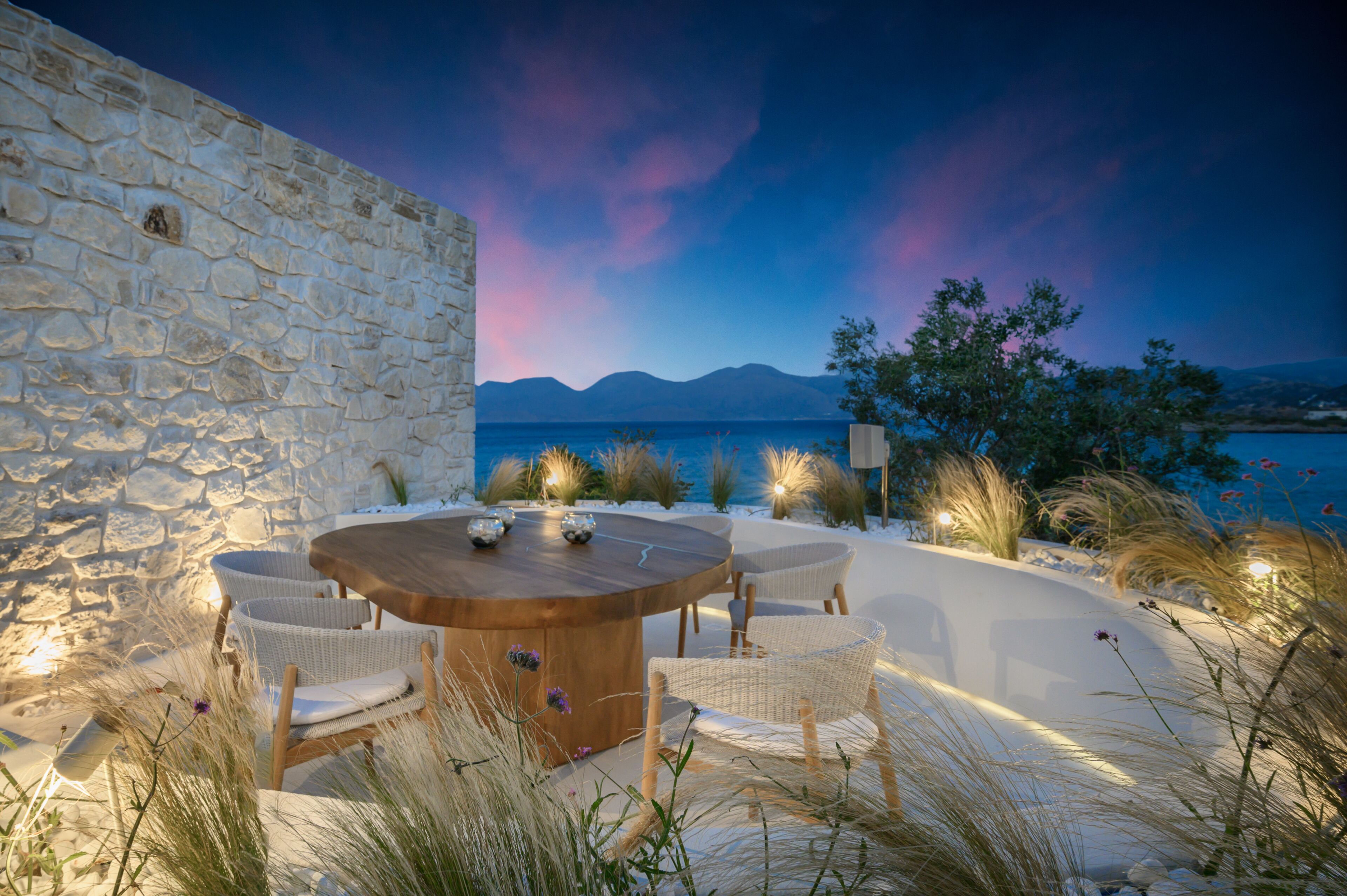 A stone wall, a round wooden table with chairs, and a view of the sea and mountains.