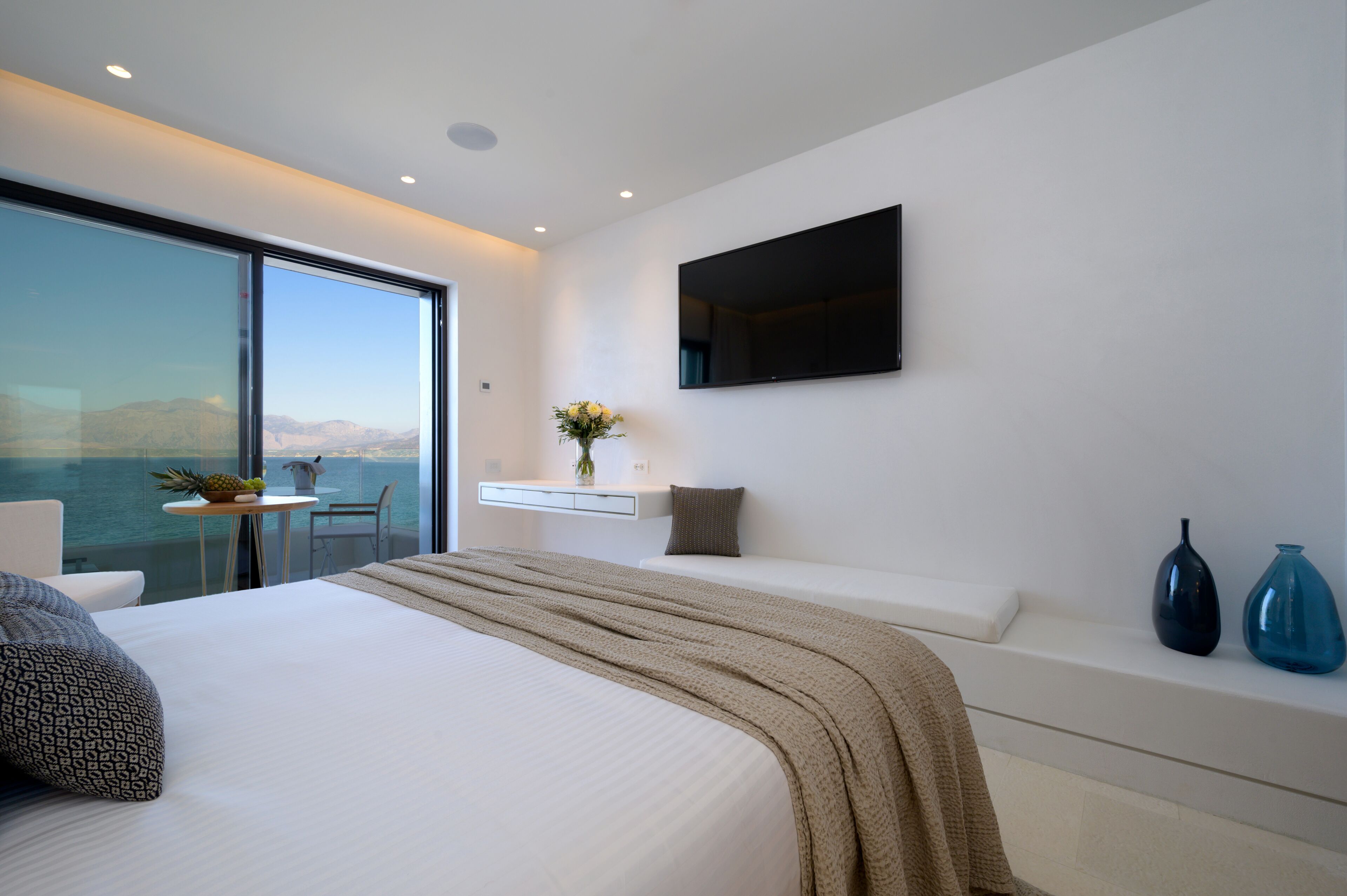 A modern bedroom with a large bed, a TV mounted on the wall, a small table with a vase of flowers, and a view of the sea through a large window.