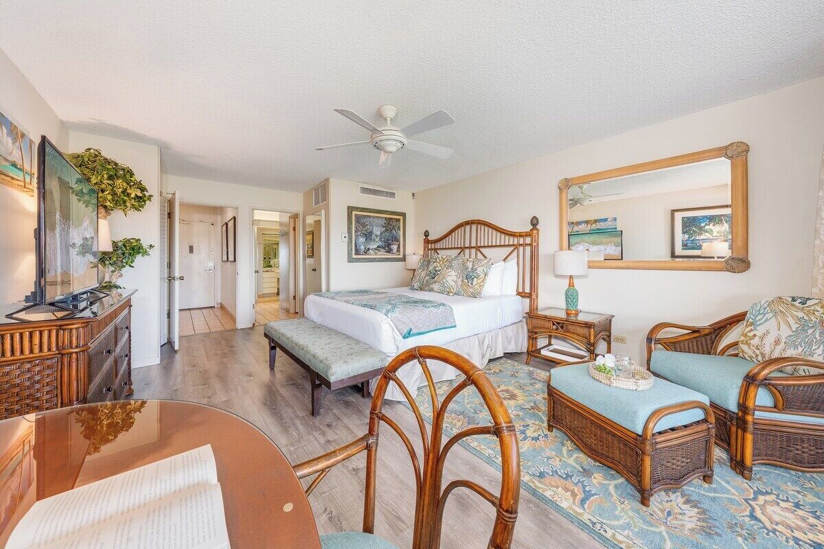 Une chambre d’hôtel avec un lit, deux fauteuils en osier, une petite table, un miroir et un ventilateur de plafond.